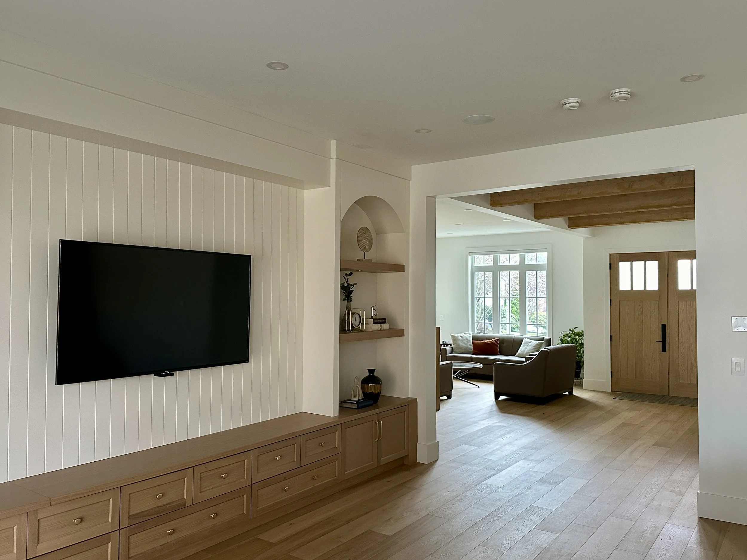 Living room with flat-screen TV on white paneled wall, wooden sideboard, open shelving with decorative items, and a sitting area with sofa and armchairs near large windows, wooden ceiling beams, and a wooden front door.