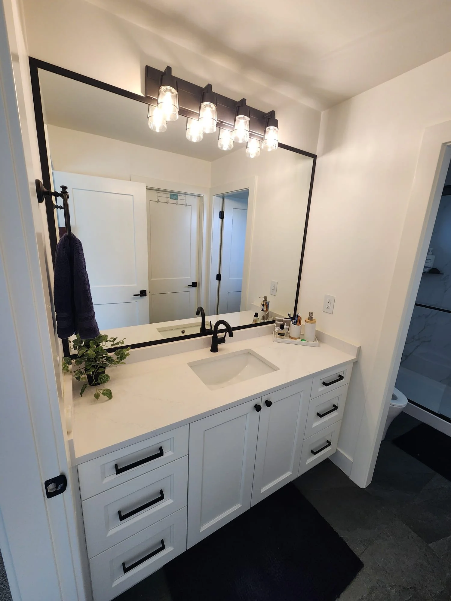 Modern white bathroom vanity with a large mirror, black fixtures, and a decorative tray with bottles, next to a small plant, with a laundry area visible in the background.