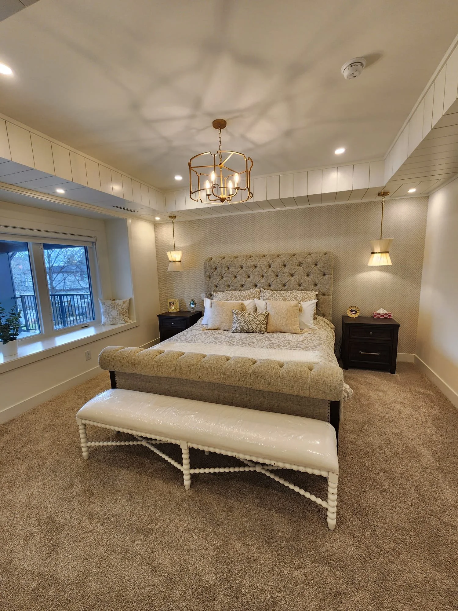 A bedroom with a large bed with beige tufted headboard, multiple decorative pillows, dark nightstands with lamps, a bench at the foot of the bed, a window with a view outside, and ceiling lighting fixtures.