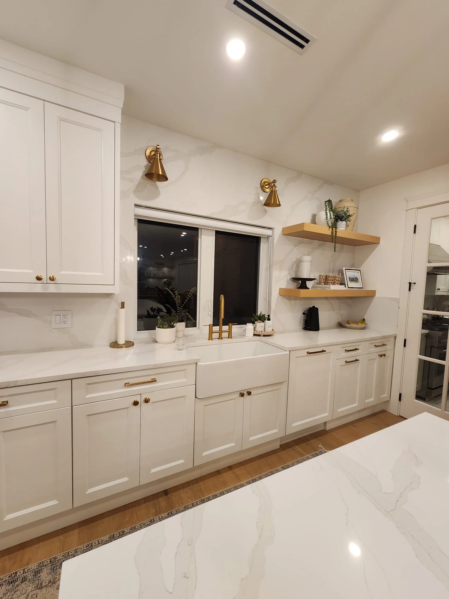 Modern kitchen with white cabinets, marble countertops, and gold fixtures. There are two open wooden shelves with decor items, a window above the sink, and a door to the right. The space is well-lit with recessed lighting.