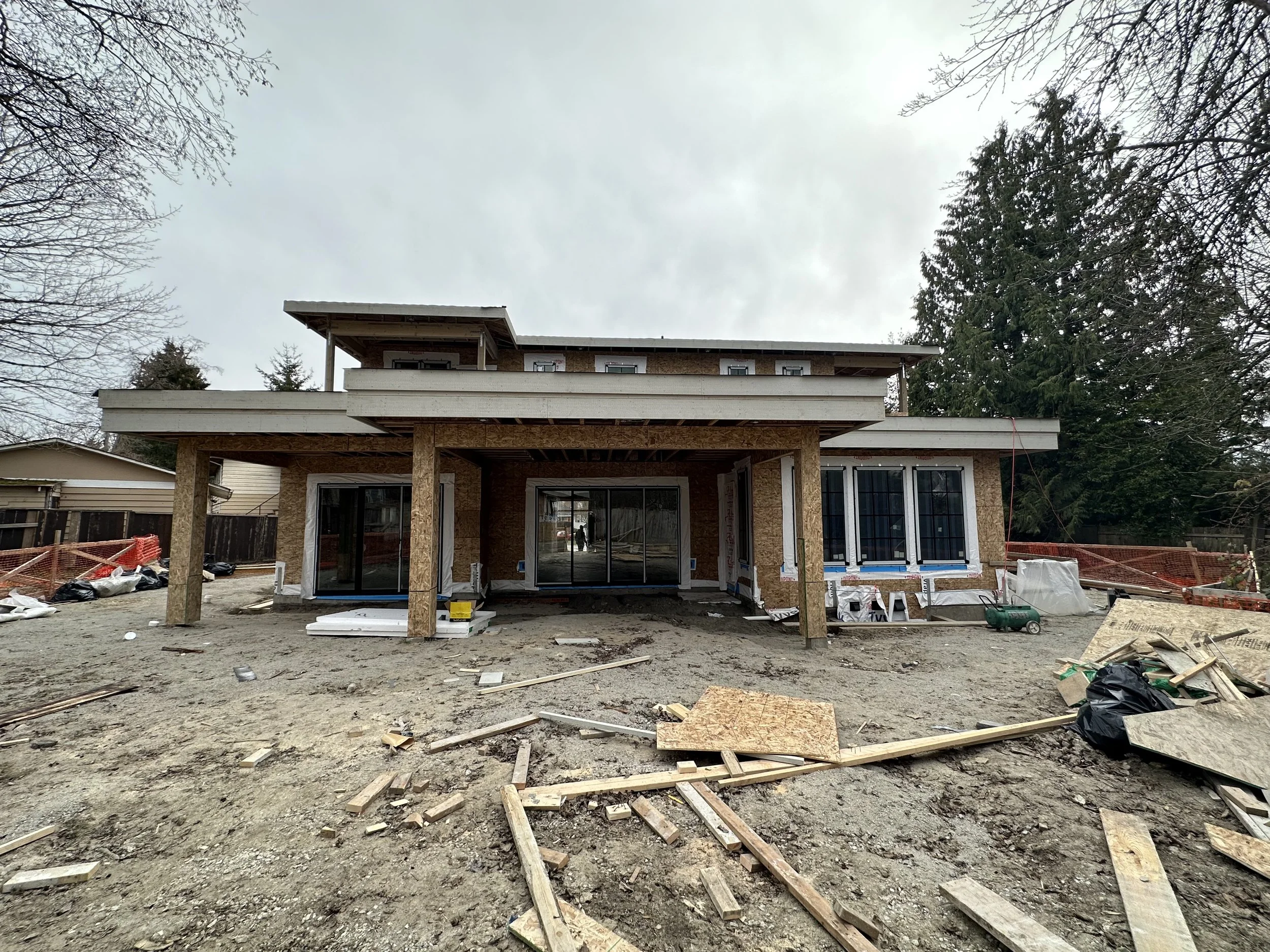 Under construction house with exposed wooden framing, large sliding glass doors, and construction materials scattered in the front yard on a cloudy day.