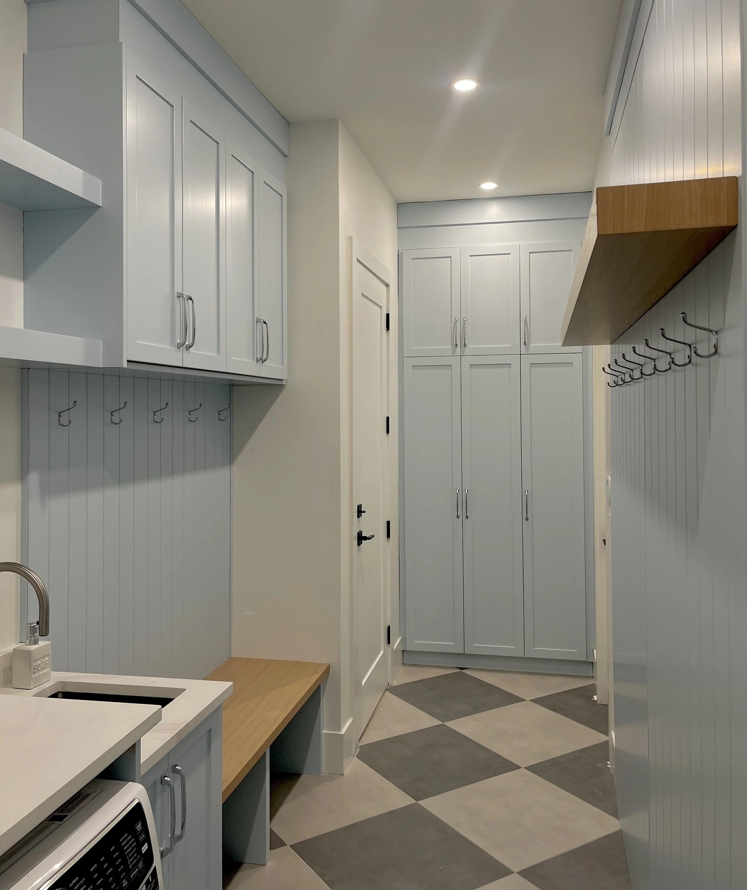 A laundry or utility room with white cabinets, a wooden shelf, hooks on the wall, a bench, a washing machine, and checkered black and beige tile flooring.