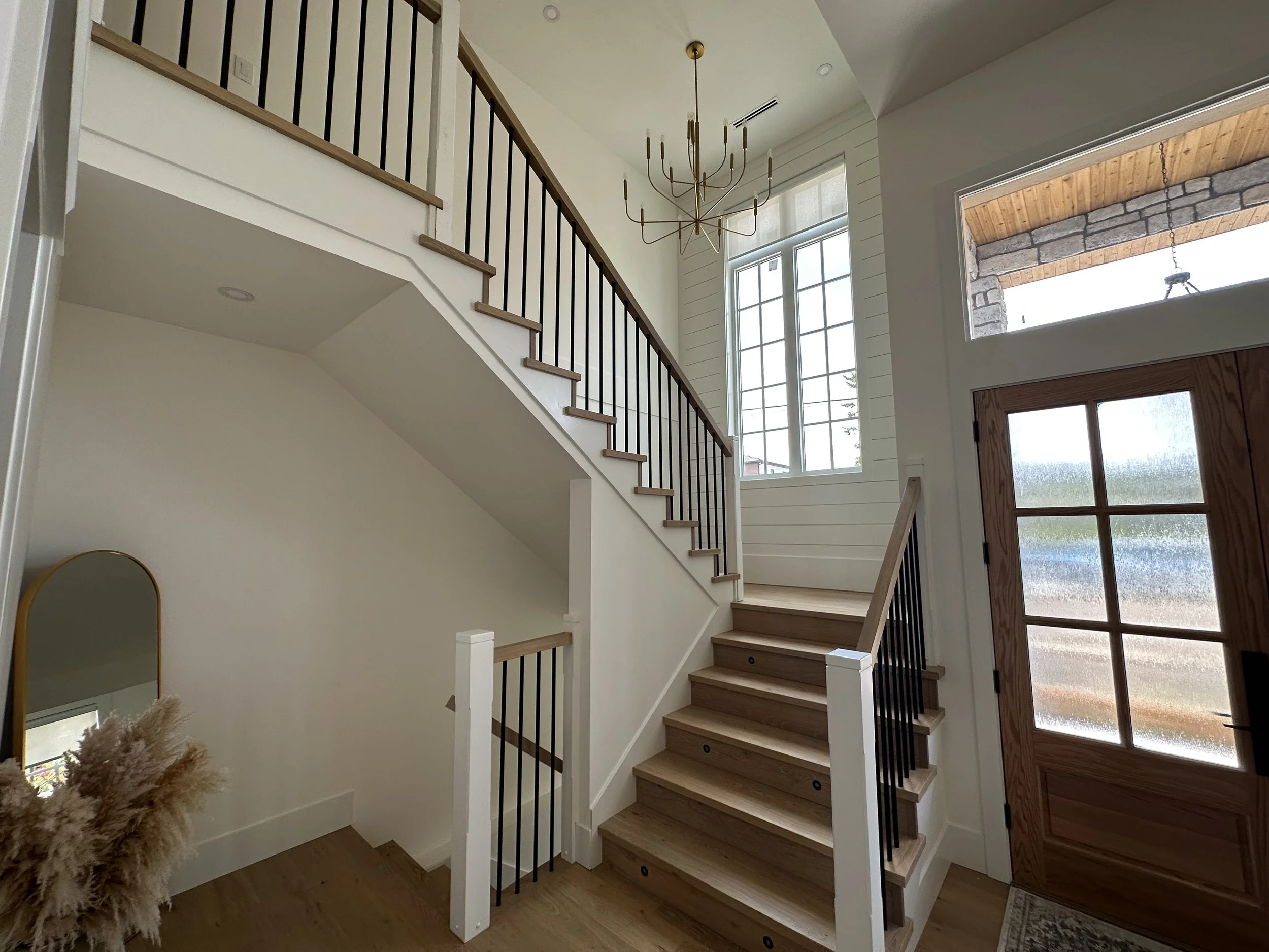 Interior view of a staircase with wooden steps, black metal railing, and large windows behind. A pendant chandelier hangs from the ceiling, and a wooden front door with a window is visible to the right.