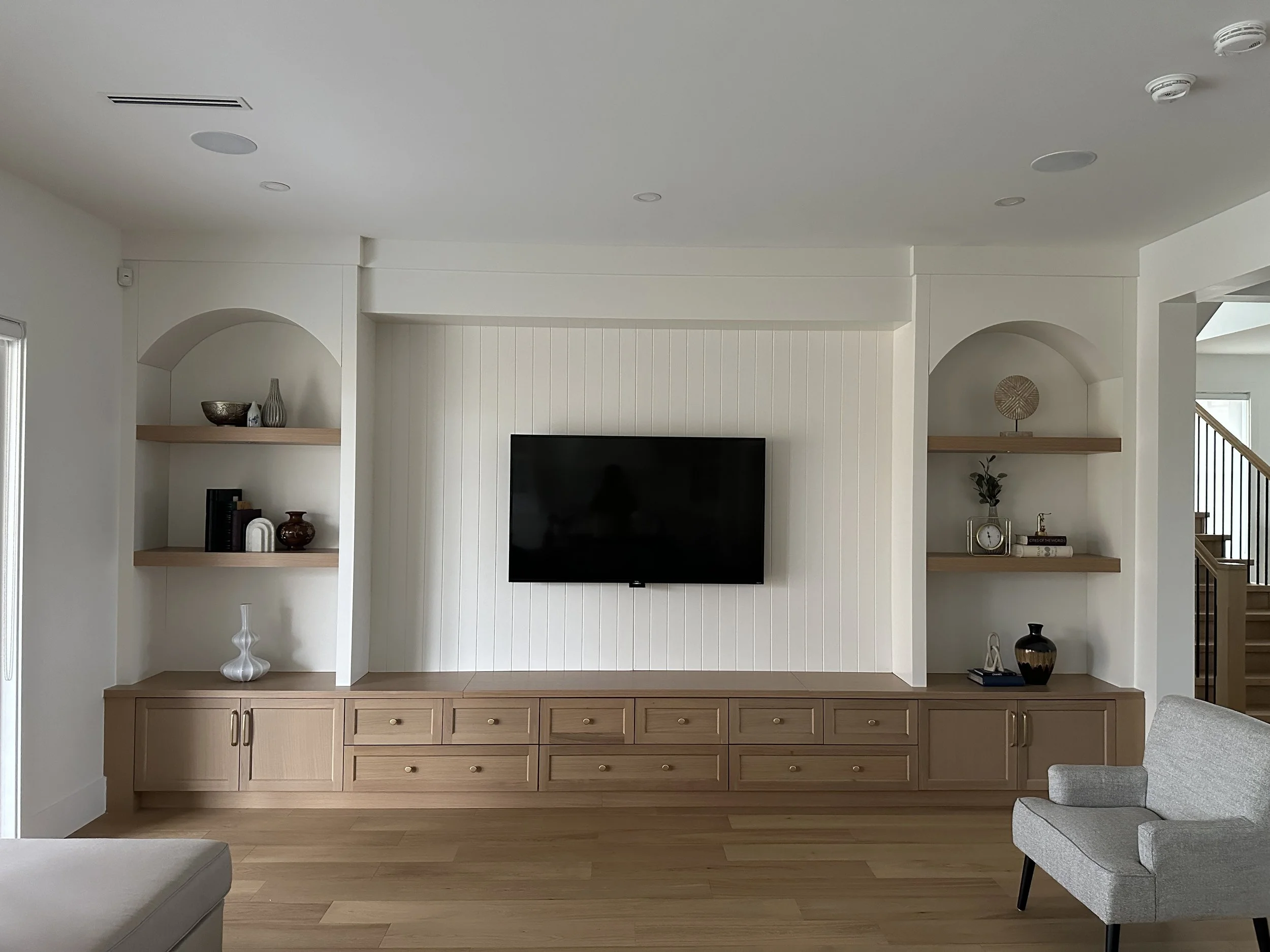 Living room with built-in wooden media console, white shelves with decorative items, and a flat-screen TV mounted on a white paneled wall. Part of a gray armchair and a staircase are visible.