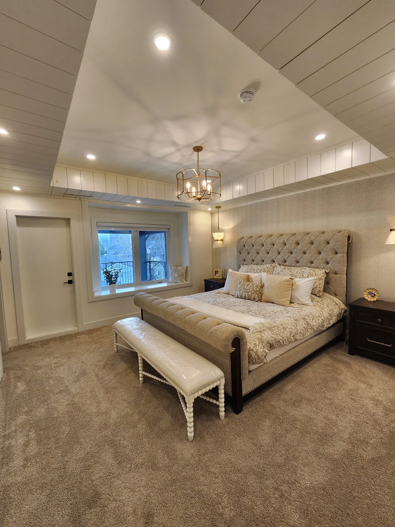 A cozy bedroom with beige walls, carpet, and a large bed with a tufted headboard, decorative pillows, and a matching bench at the foot of the bed. There are dark nightstands on each side of the bed, a window with a potted plant, and ceiling lighting