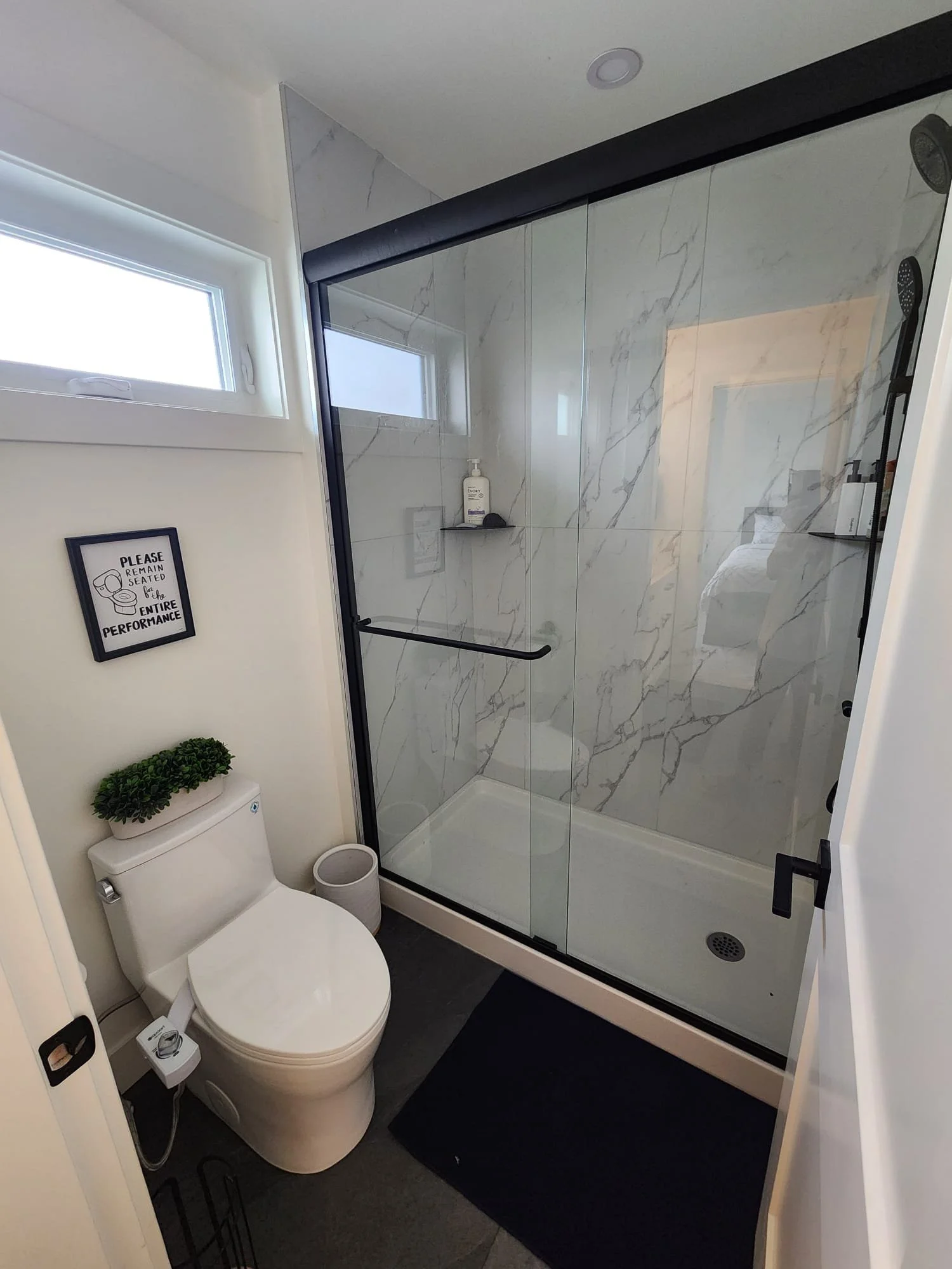 A bathroom with a glass shower enclosure, a toilet with a small plant on top, a framed sign on the wall, a small window, and a black rug on the gray floor.