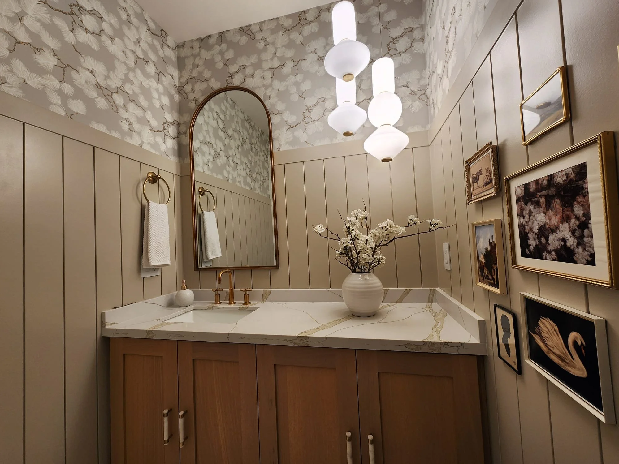 A bathroom with beige paneling, floral wallpaper, a marble countertop with a vessel sink, a gold faucet, a large standing mirror, wall-mounted light fixtures, a vase with white flowers, a gallery wall of framed pictures, and a white towel on a gold r