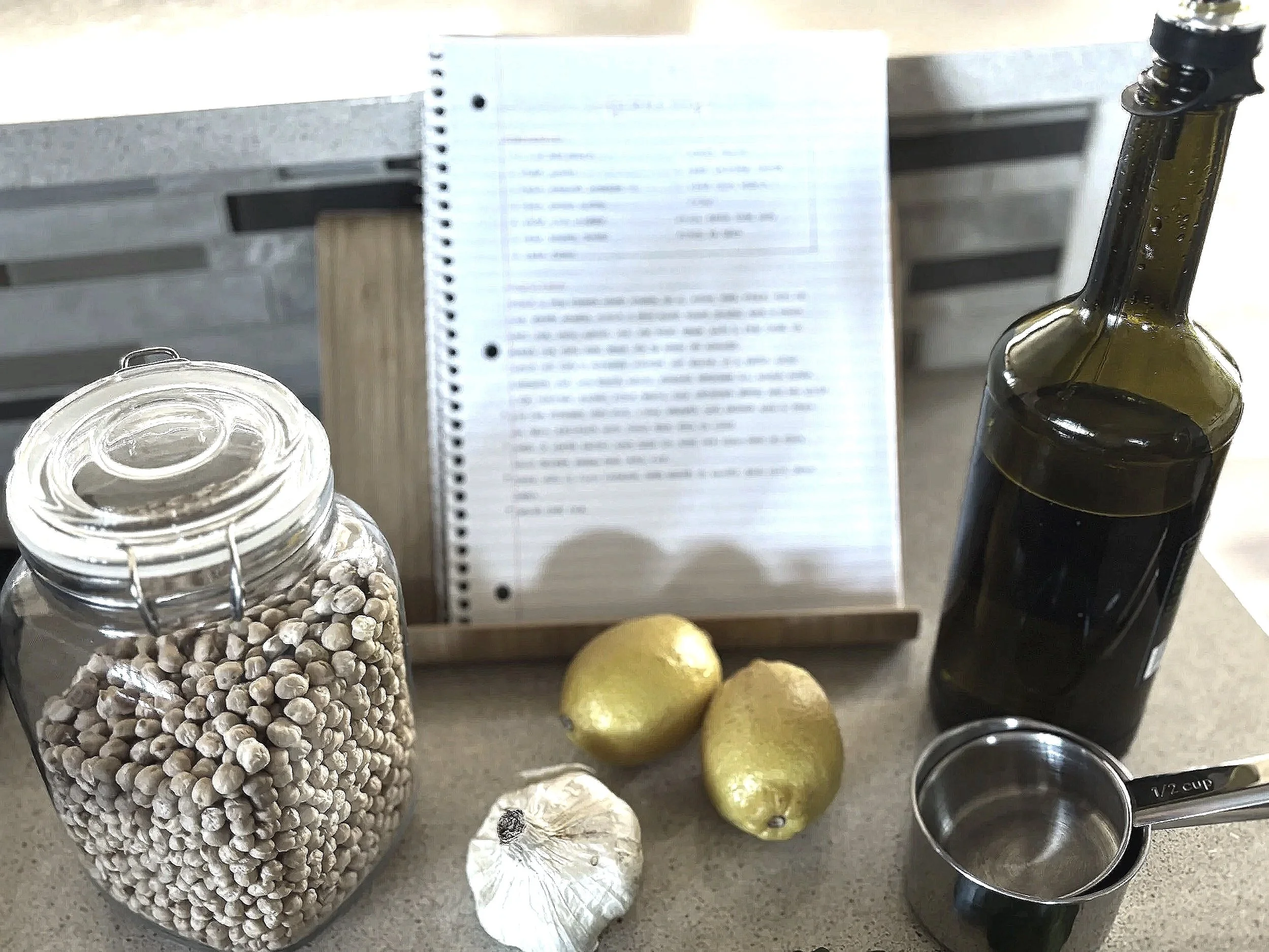 A kitchen countertop with a jar of chickpeas, two lemons, a head of garlic, a bottle of olive oil, a measuring cup, and a handwritten recipe in a wooden stand.