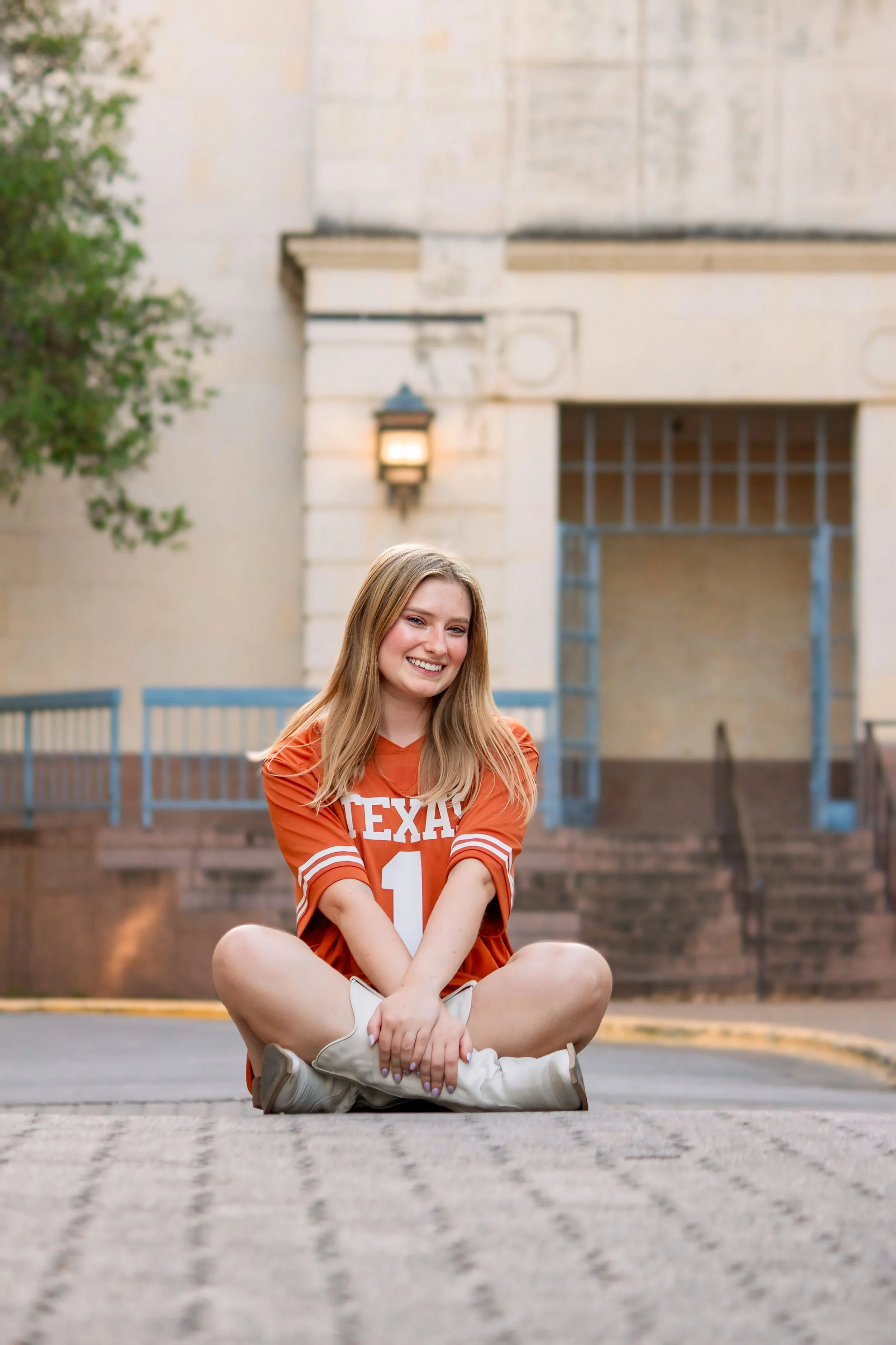 Young woman with blonde hair sitting cross-legged on the pavement in a Texas football jersey, smiling at the camera, with a building and stairs in the background.