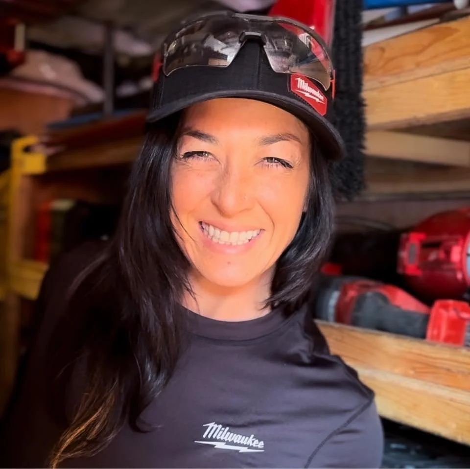 Woman with long dark hair smiling, wearing a black Milwaukee cap with safety glasses on top, in a workshop with power tools and wooden shelves in the background.