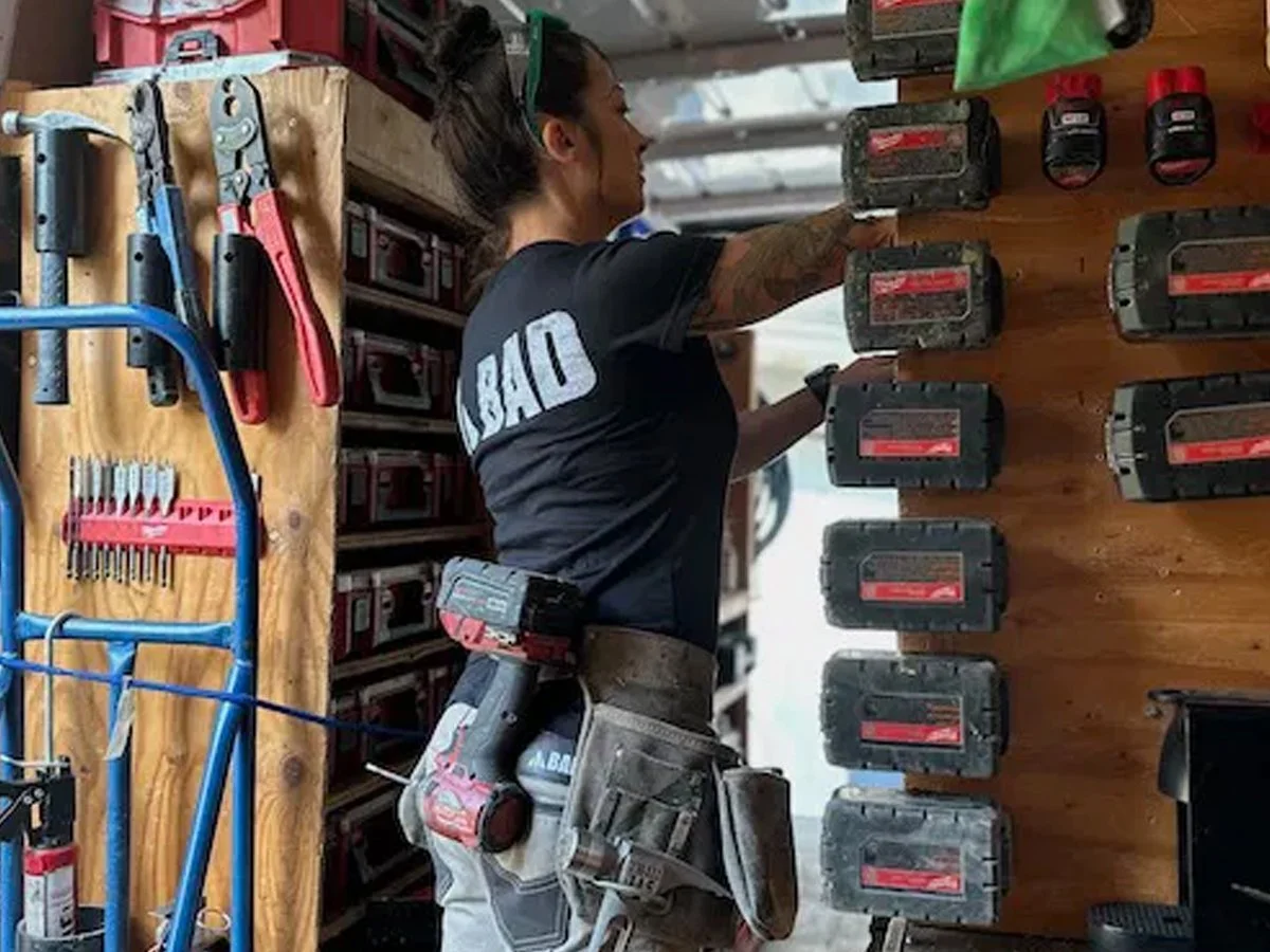A woman working on a wooden wall with power tools organized on a pegboard and shelf, wearing a black t-shirt with the word 'BAD' on the back, and a tool belt with a cordless drill
