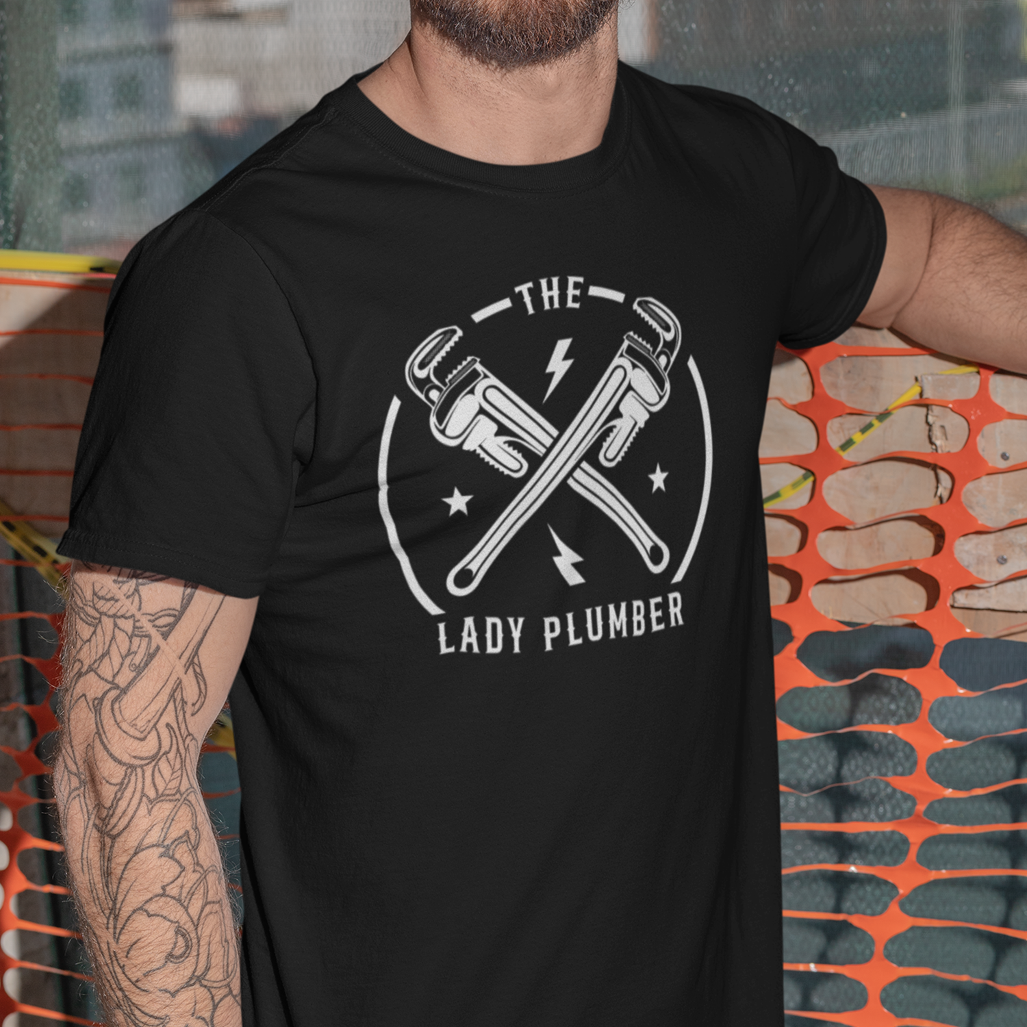 Close-up of a man wearing a black T-shirt with a graphic design of two crossed plumbing wrenches, a lightning bolt, stars, and the text 'The Lady Plumber' on it.