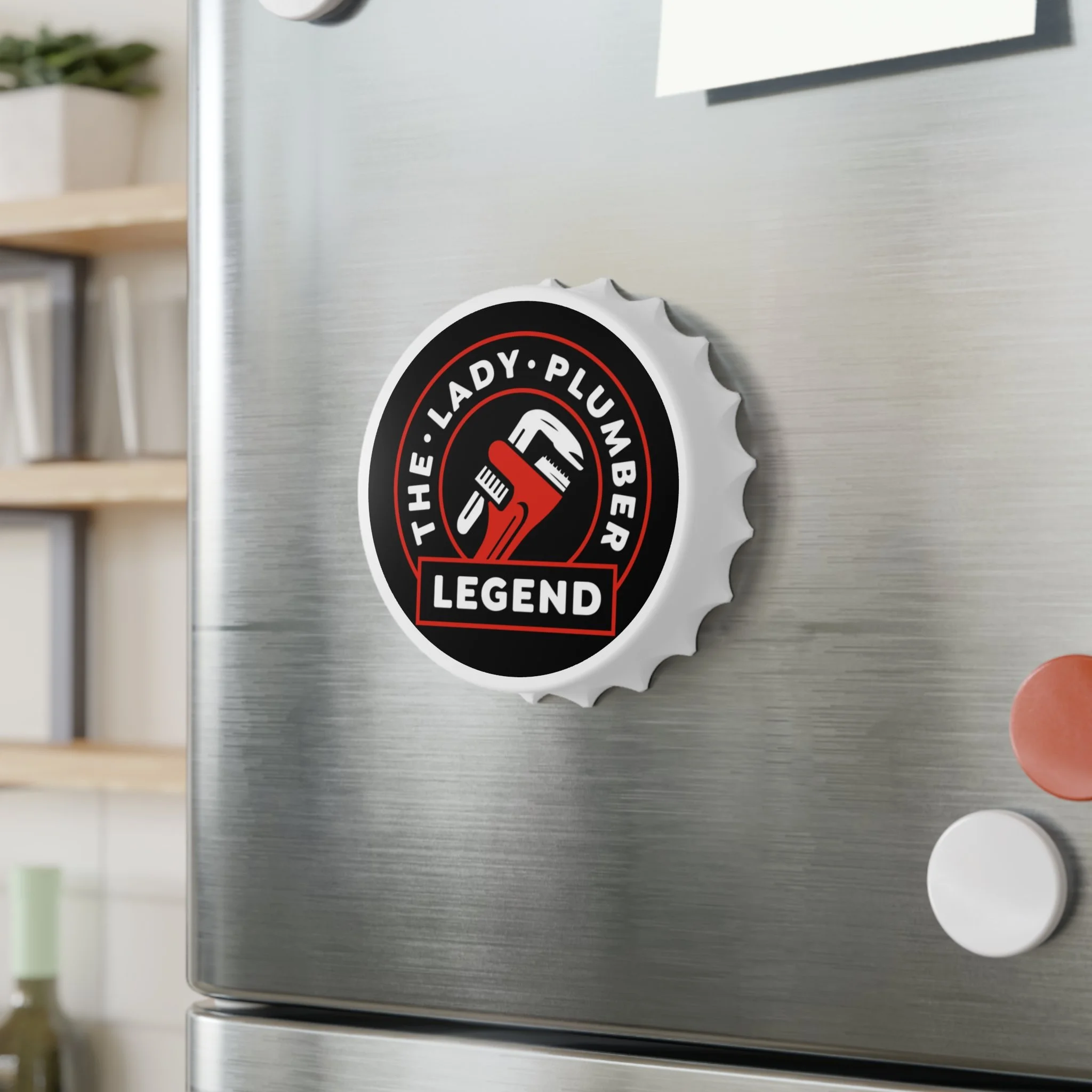 Stainless steel refrigerator door with a round magnet that has a black, white, and red design. The magnet features a wrench and screwdriver crossed with a label that reads "The Lady-Plumber Legend."