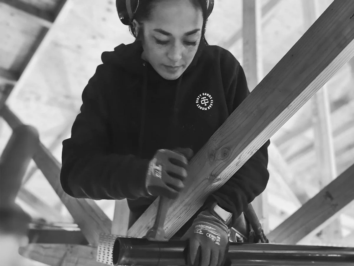A woman wearing a hoodie, gloves, and headphones is working with a hammer and a metal pipe on a wooden structure.