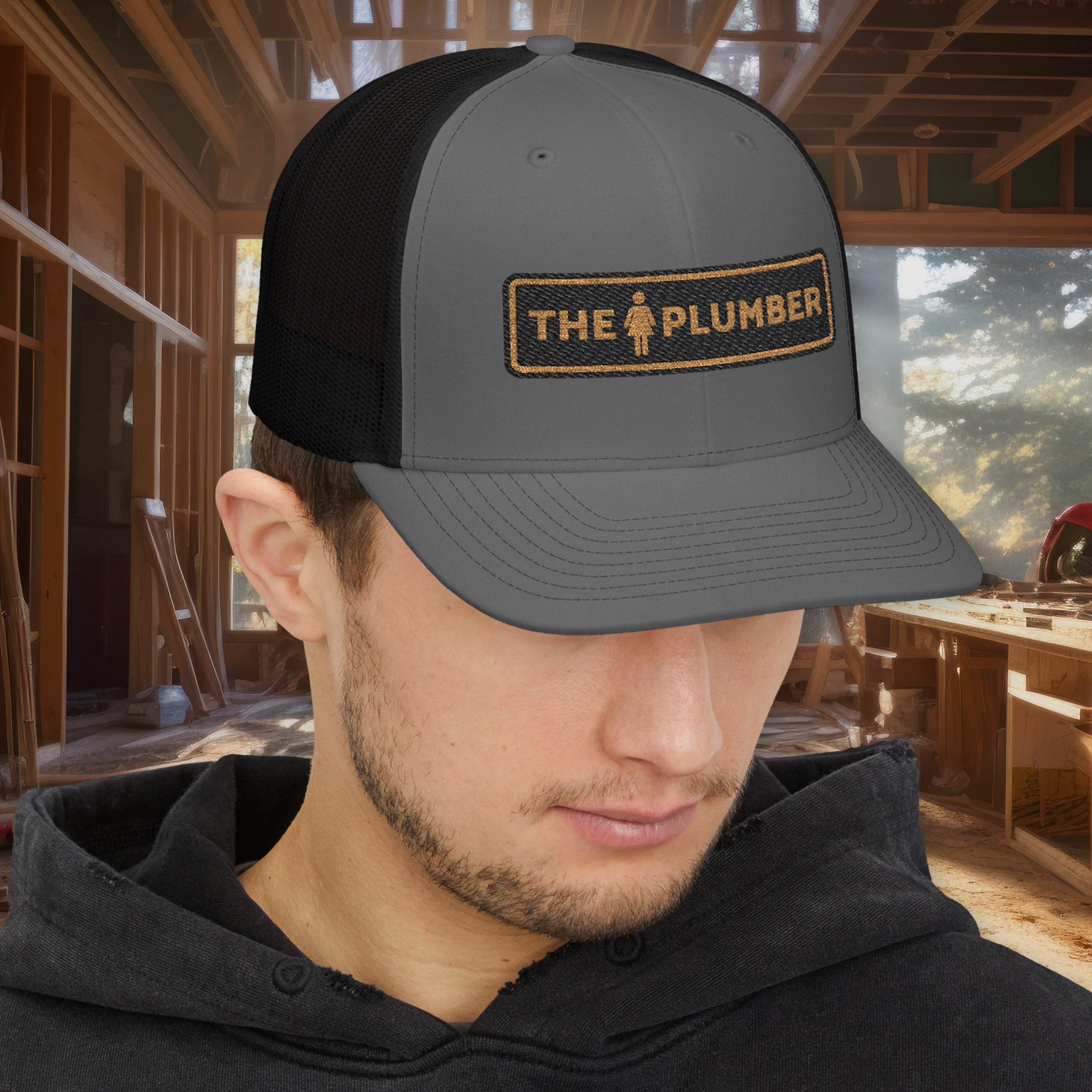 A man wearing a gray and black trucker cap with an embroidered patch that says "The Plumber" and has a female figure icon. The background features an unfinished wood interior with large windows showing trees and a sky.