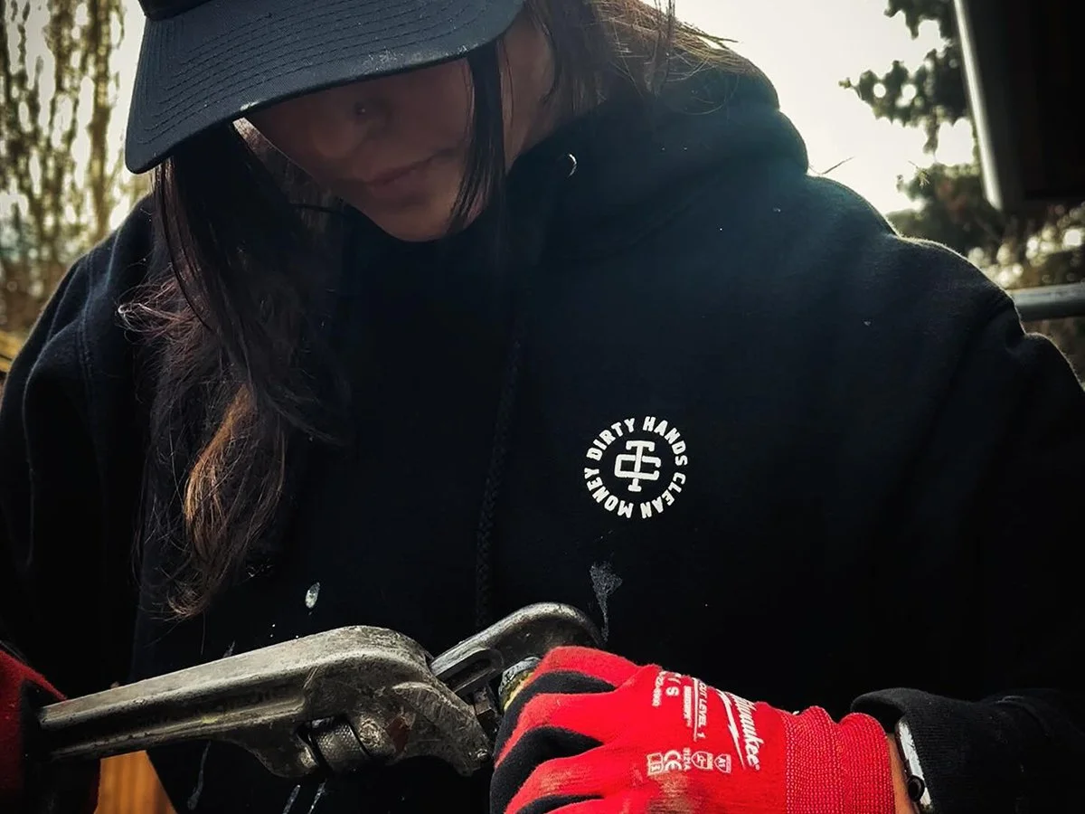 Person wearing a black hoodie with a logo, a gray cap, red work gloves, and using a wrench outdoors.