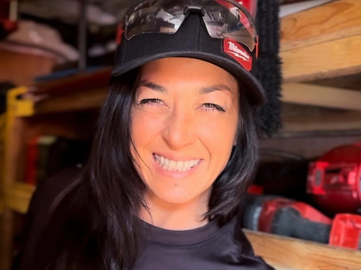 A woman smiling, wearing a black hat with a Milwaukee tool logo, sunglasses on her hat, and a black shirt, standing in a workshop or tool storage area with wooden shelves and red power tools in the background.