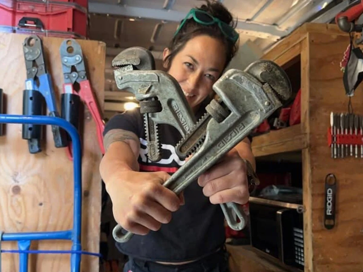 A woman in a workshop holding two large pipe wrenches and smiling at the camera. Behind her are various tools hanging on a wooden wall.