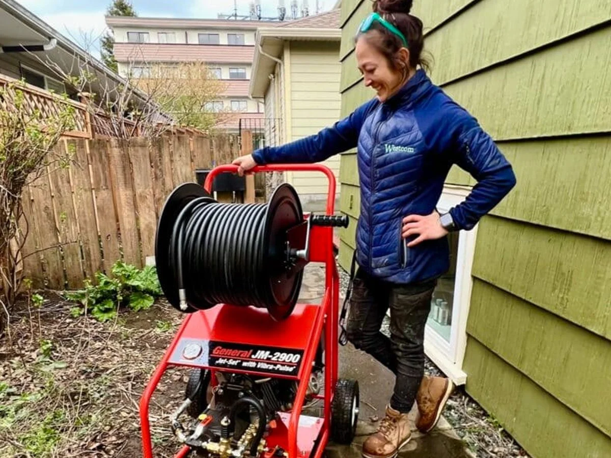 A woman in outdoor work gear, with a blue jacket, camo pants, and brown work boots, smiling while holding a hose reel on a red cart or machine outside a house with green siding.