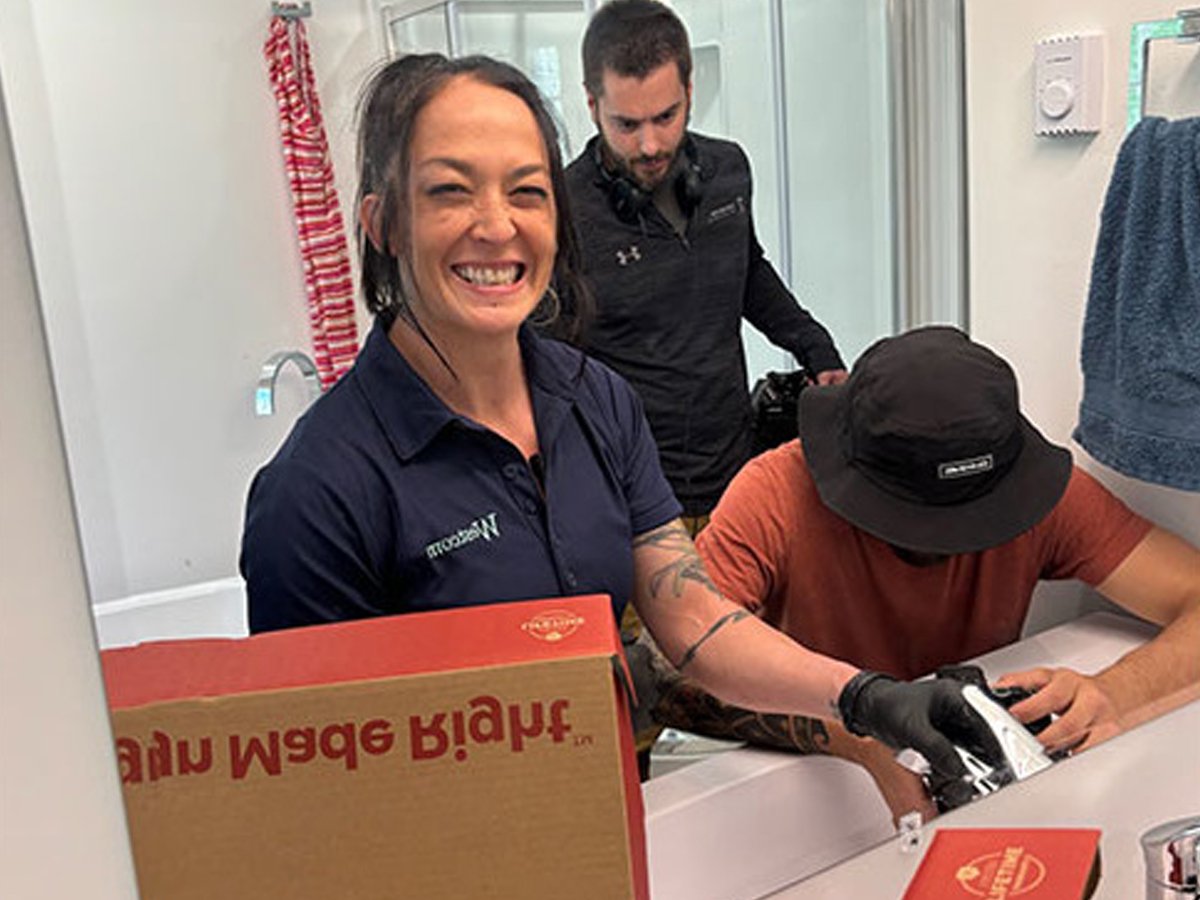 A woman smiling and showing her teeth at a restaurant counter with two men, one wearing a bucket hat and black gloves, and the other wearing a black shirt and headphones, in a casual indoor setting.