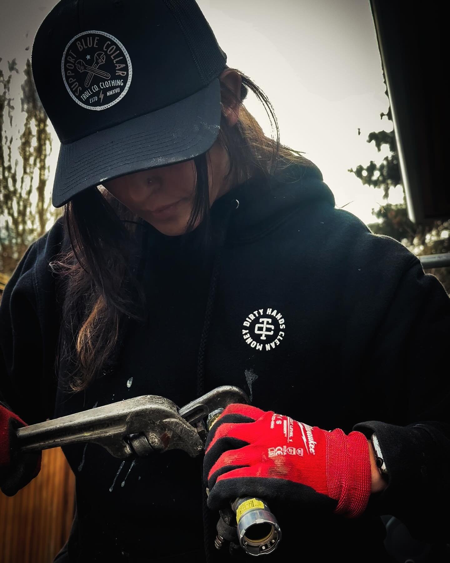 A person wearing a navy blue cap, black hoodie, and red gloves working with metal tools outdoors.