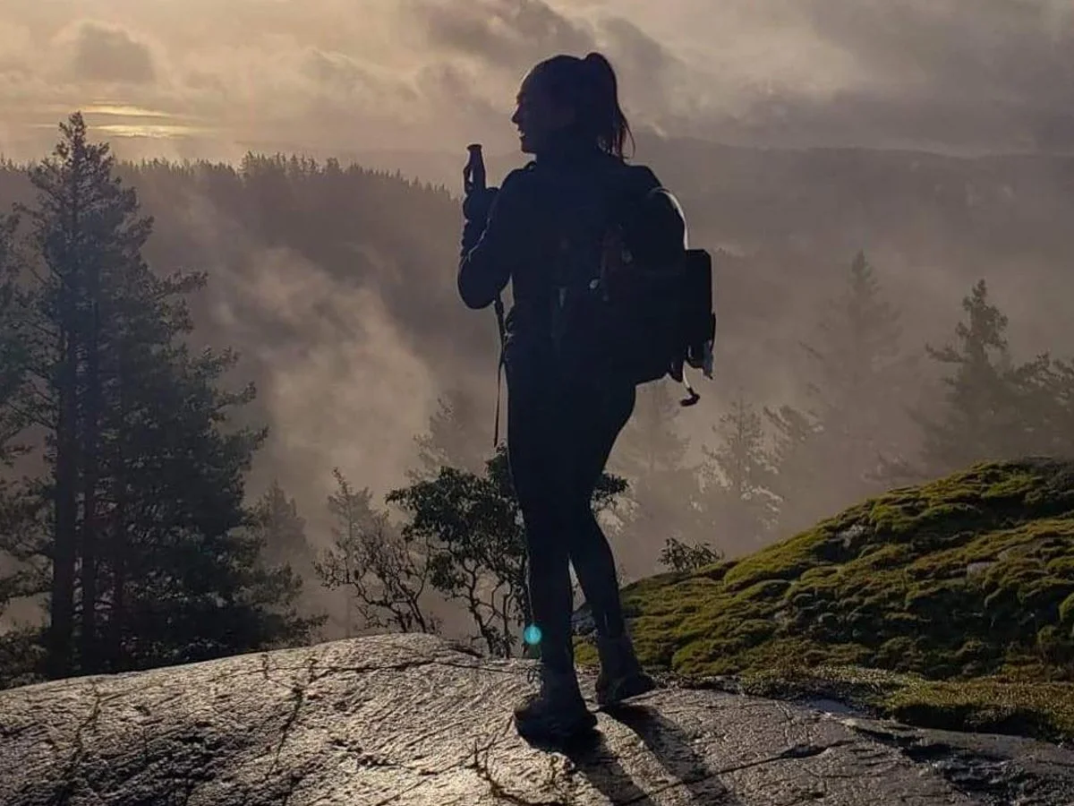 Silhouette of a woman with a backpack holding a water bottle, standing on a rocky ledge overlooking a misty forest at sunset.