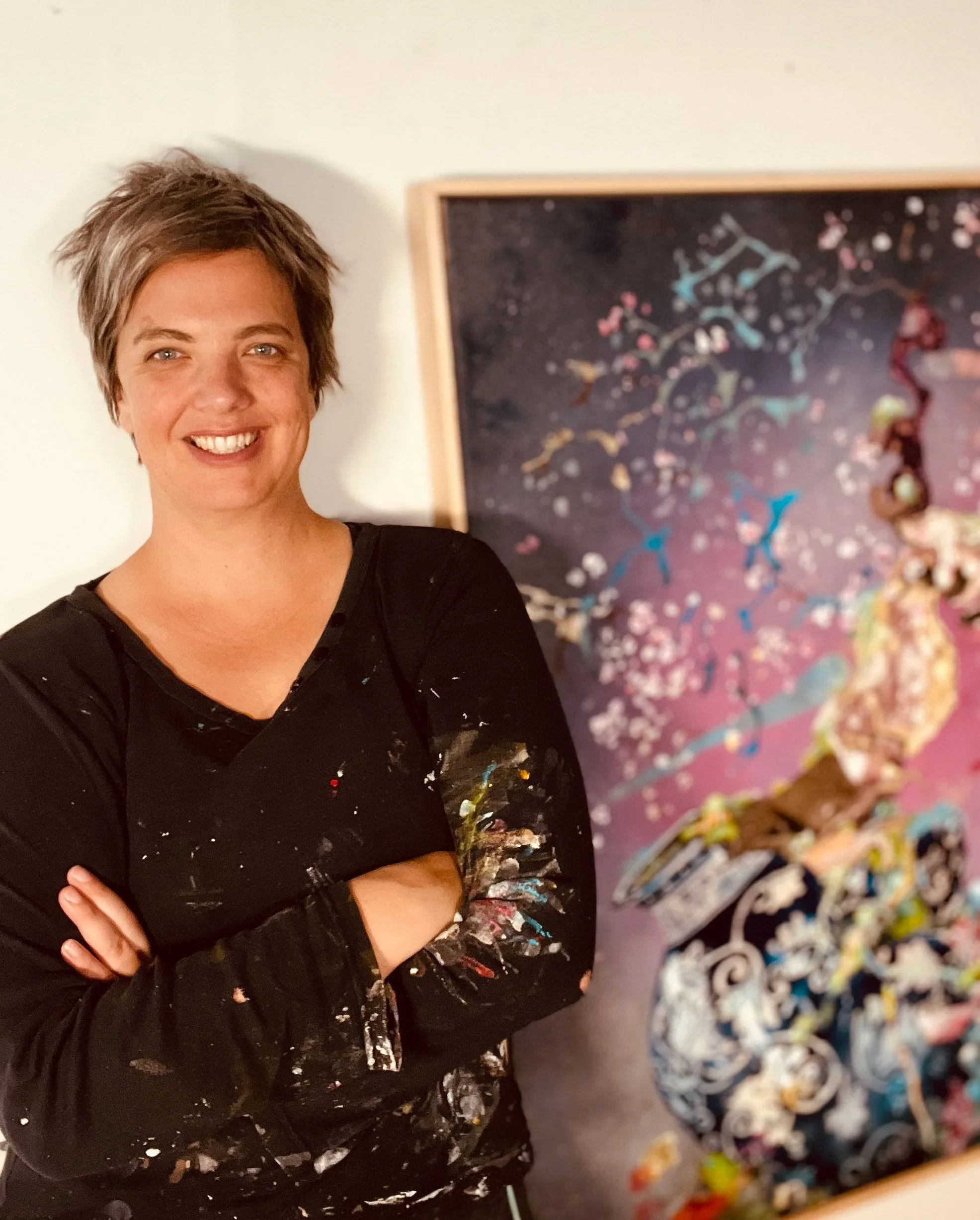 A woman with short, spiky hair and a black shirt with paint stains, smiling with arms crossed, standing in front of an abstract, colorful painting.