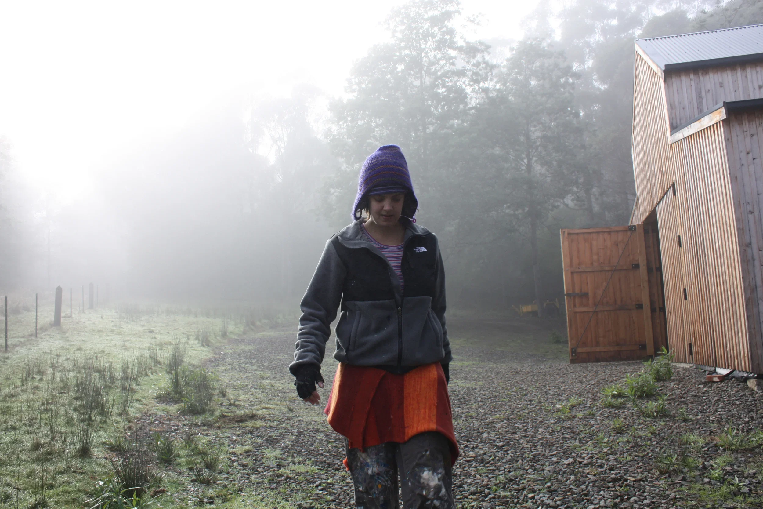 An artist walking outdoors on a foggy morning, wearing a purple knit hat, a black North Face jacket, and a red skirt. There is a wooden shed to her right used as her studio.