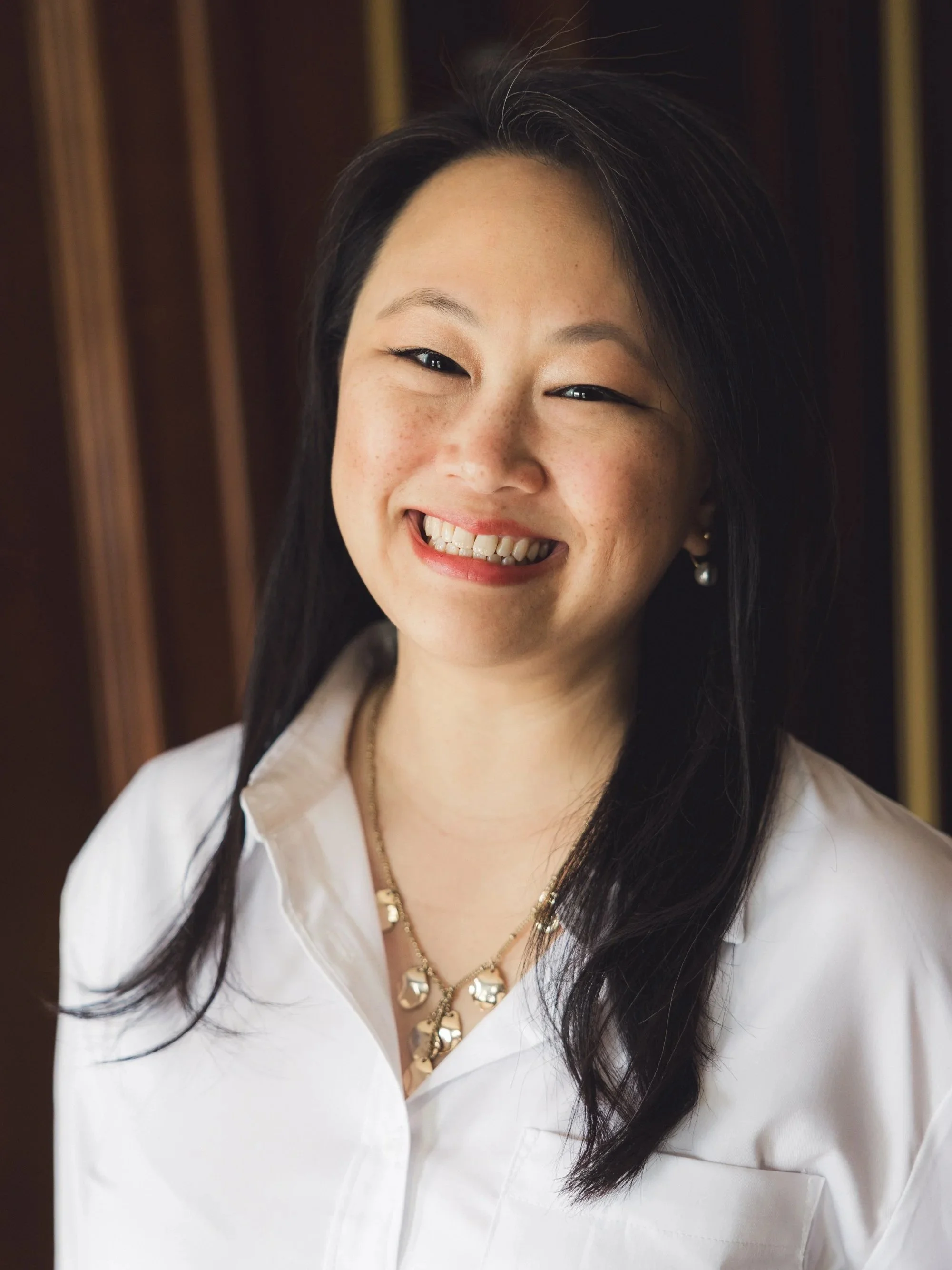 A woman with long dark hair smiling and ready support you, wearing a white blouse, gold jewelry, and earrings, against a wooden background.