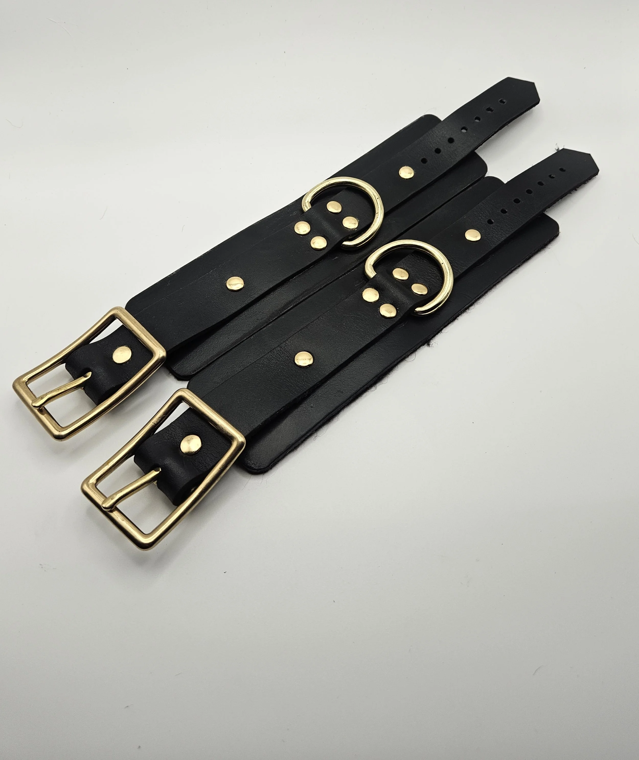 Two black leather cuffs with gold buckles and metal decorative rings, laid out on a white background.