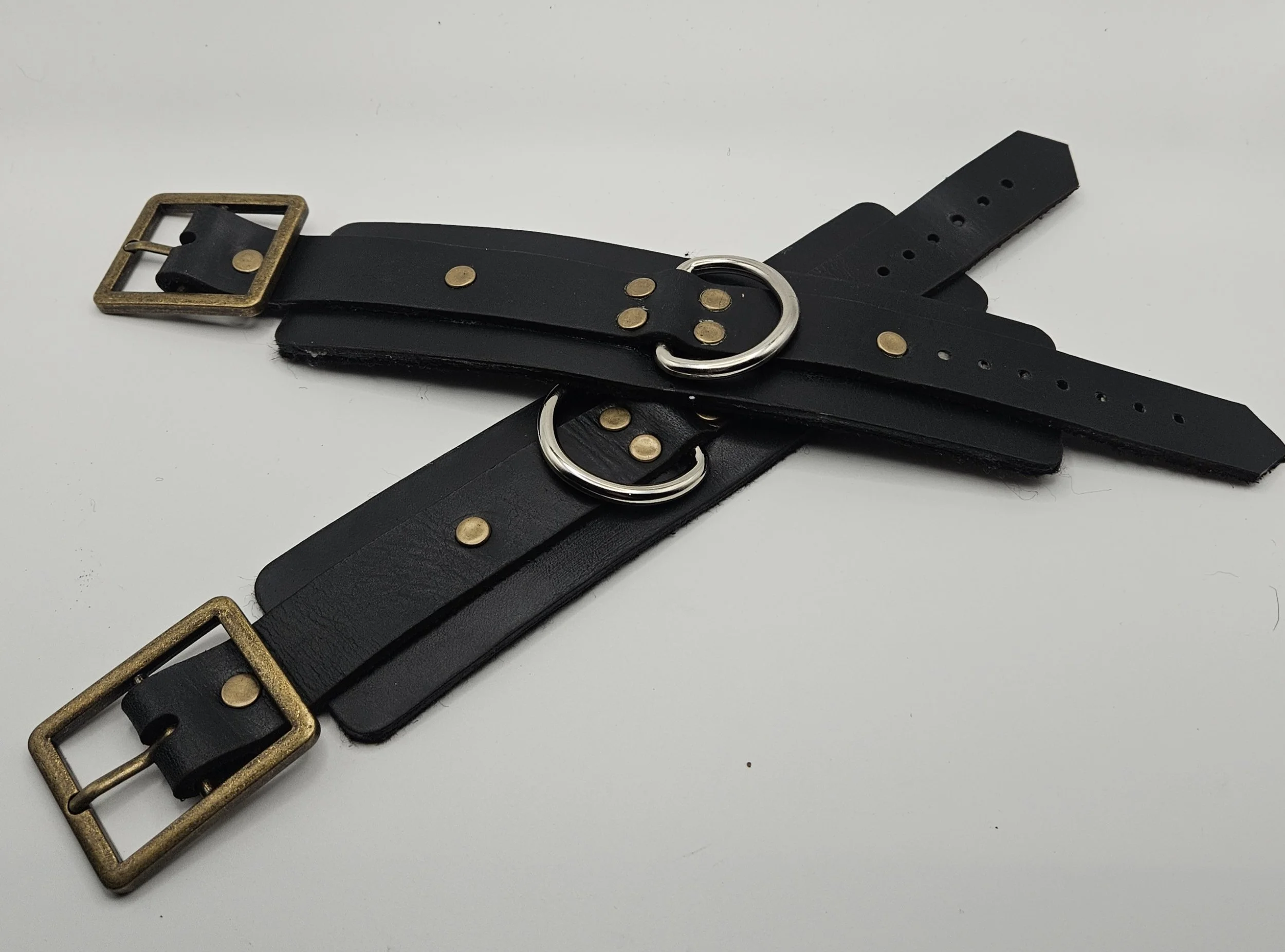Two black leather restraints with brass buckles and metal rings, intertwined and laid out on a white surface.