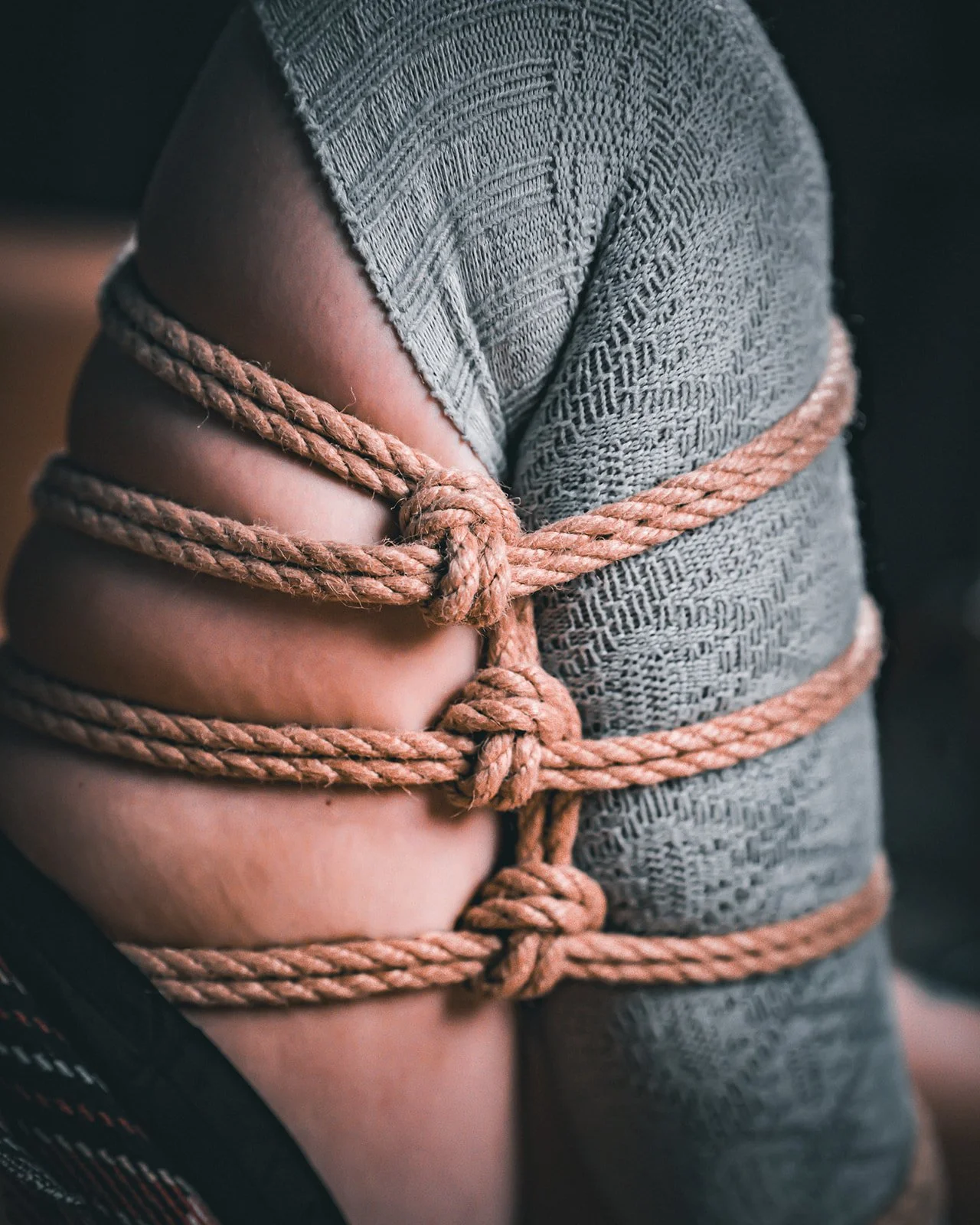 A person wrapped in multiple knots of a pinkish-brown rope, with part of their upper arm and shoulder exposed, wearing a gray patterned shirt.