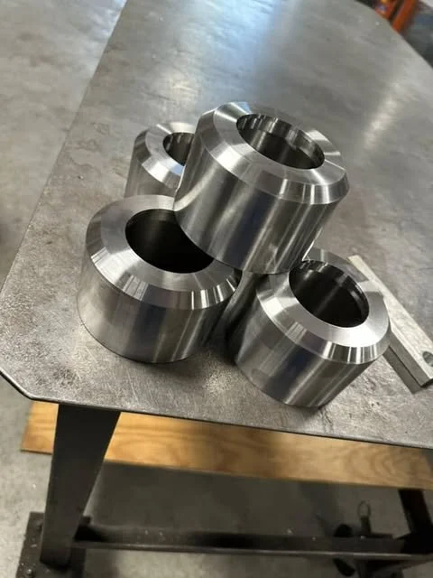 Four large, shiny metal machine parts with a cylindrical shape and a central hole, arranged on a workbench.
