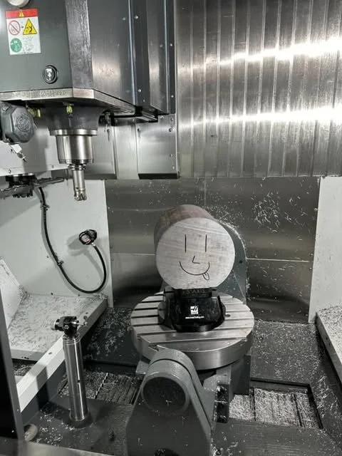A CNC machine with a wooden log on its turntable, featuring a drawn smiley face with a tongue sticking out.