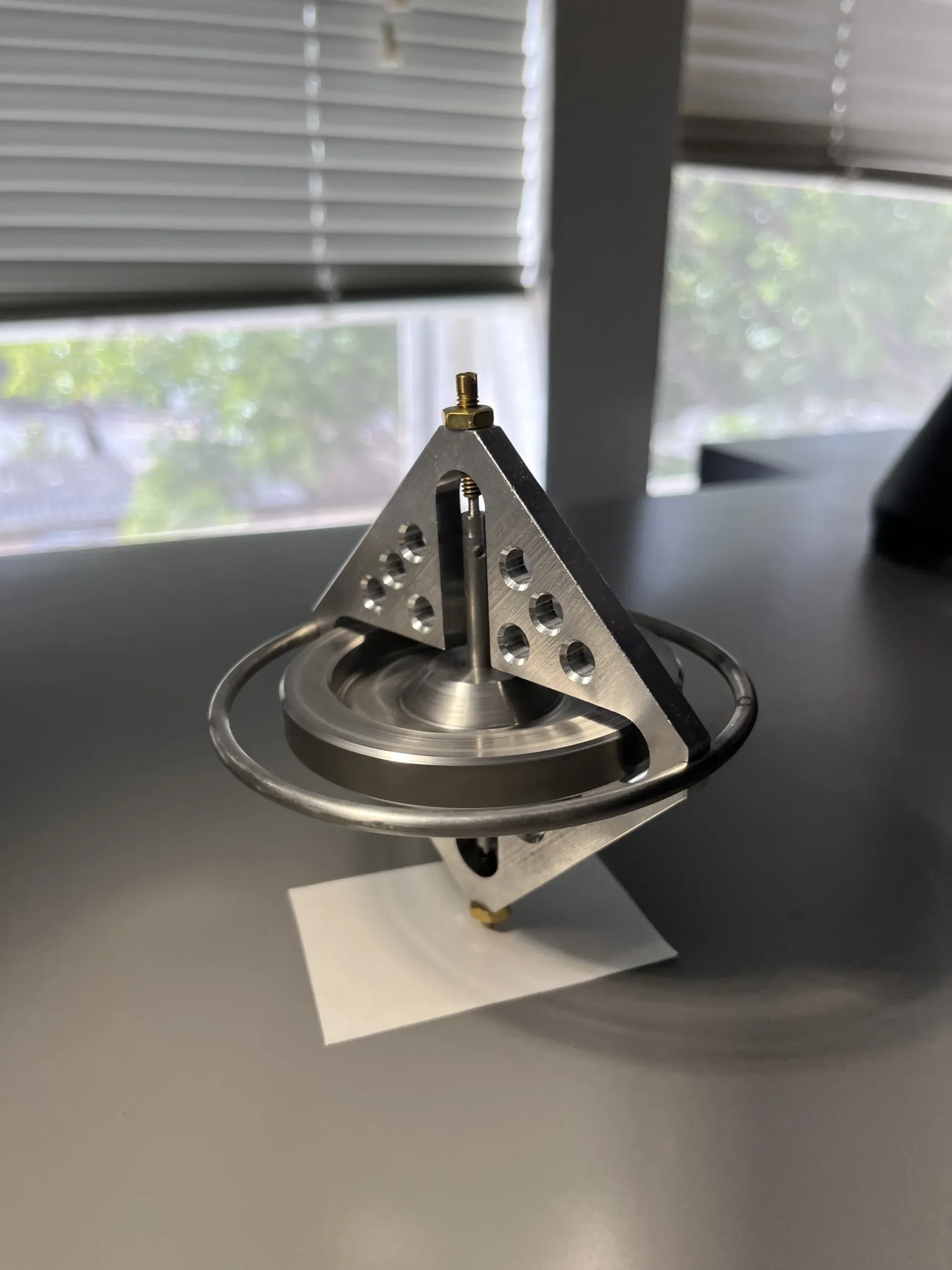 A metallic scientific or engineering device with a conical structure, circular rings, and multiple holes, placed on a white surface near a window with blinds.