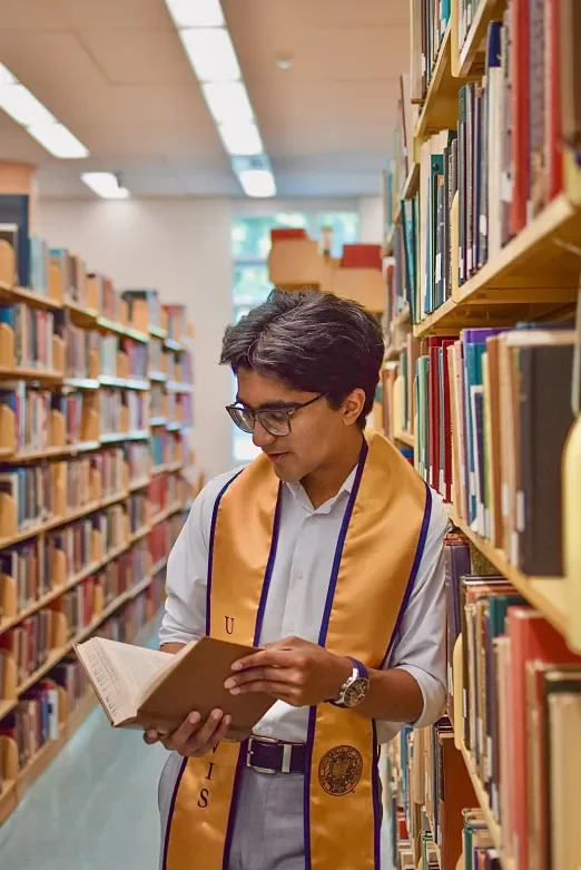Young man wearing glasses and a graduation stole reading a book in a library aisle.