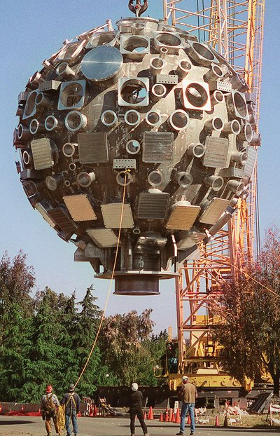 A large spherical structure covered with various electronic devices and speakers being lifted by a crane during construction.