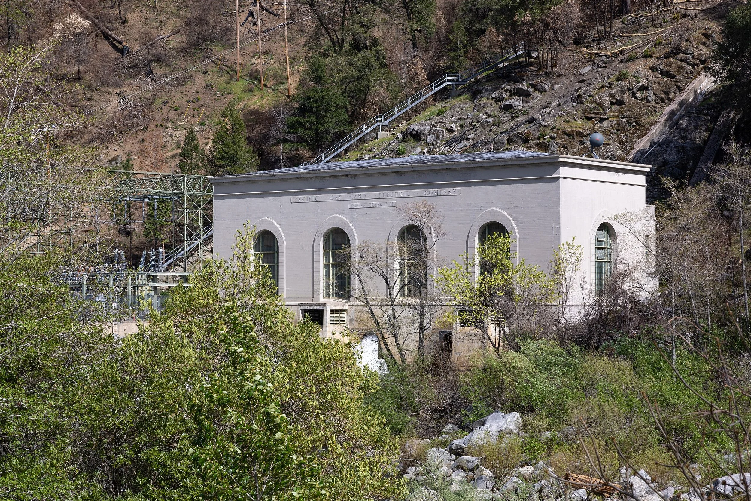 An old white building in a mountainous area with trees and rocky terrain. The building has arched windows and exterior stairs, with electrical equipment nearby.