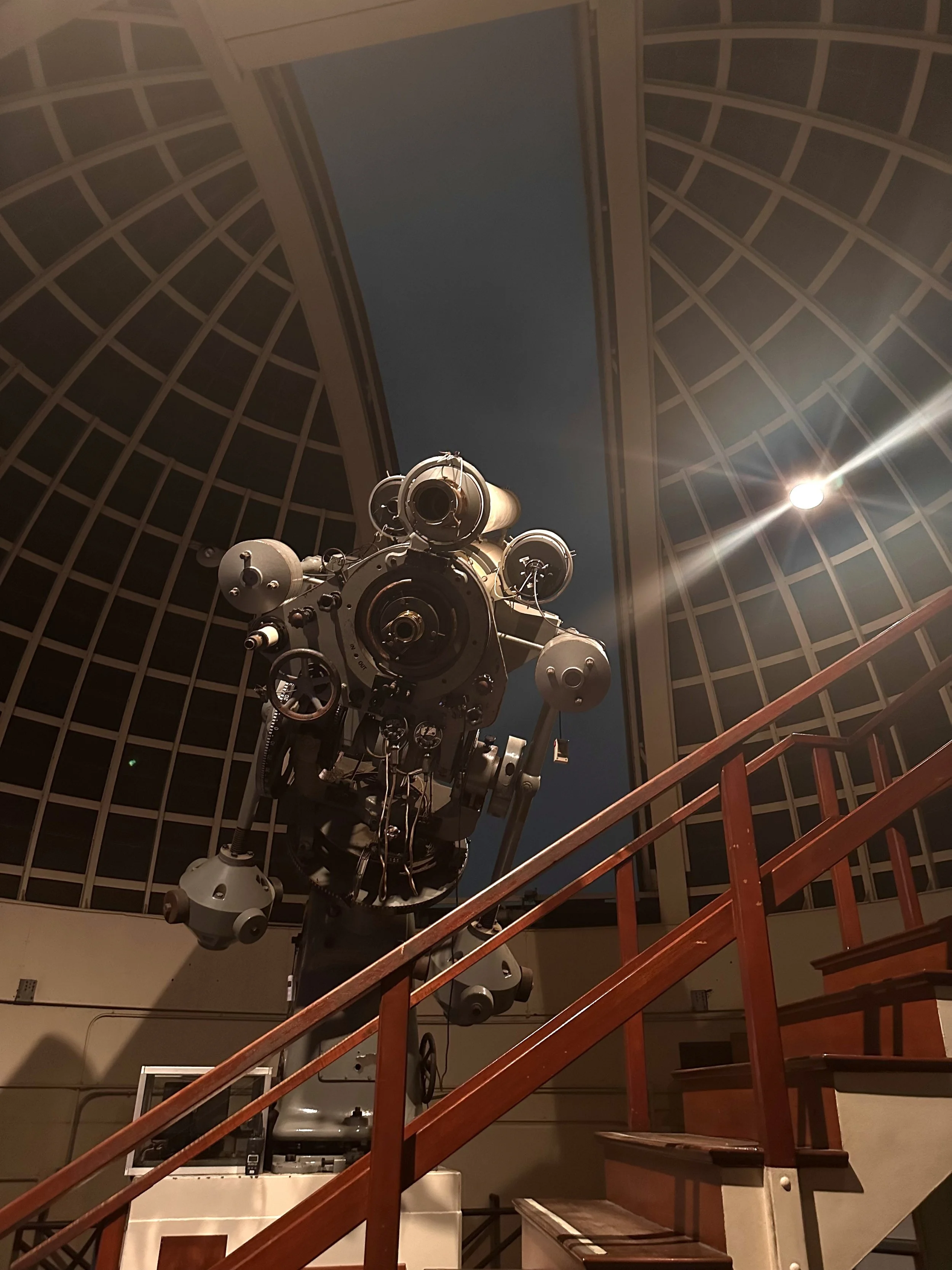 A large telescope inside an observatory, seen from below, with a staircase leading up to it. The observatory has a domed ceiling with window panels.