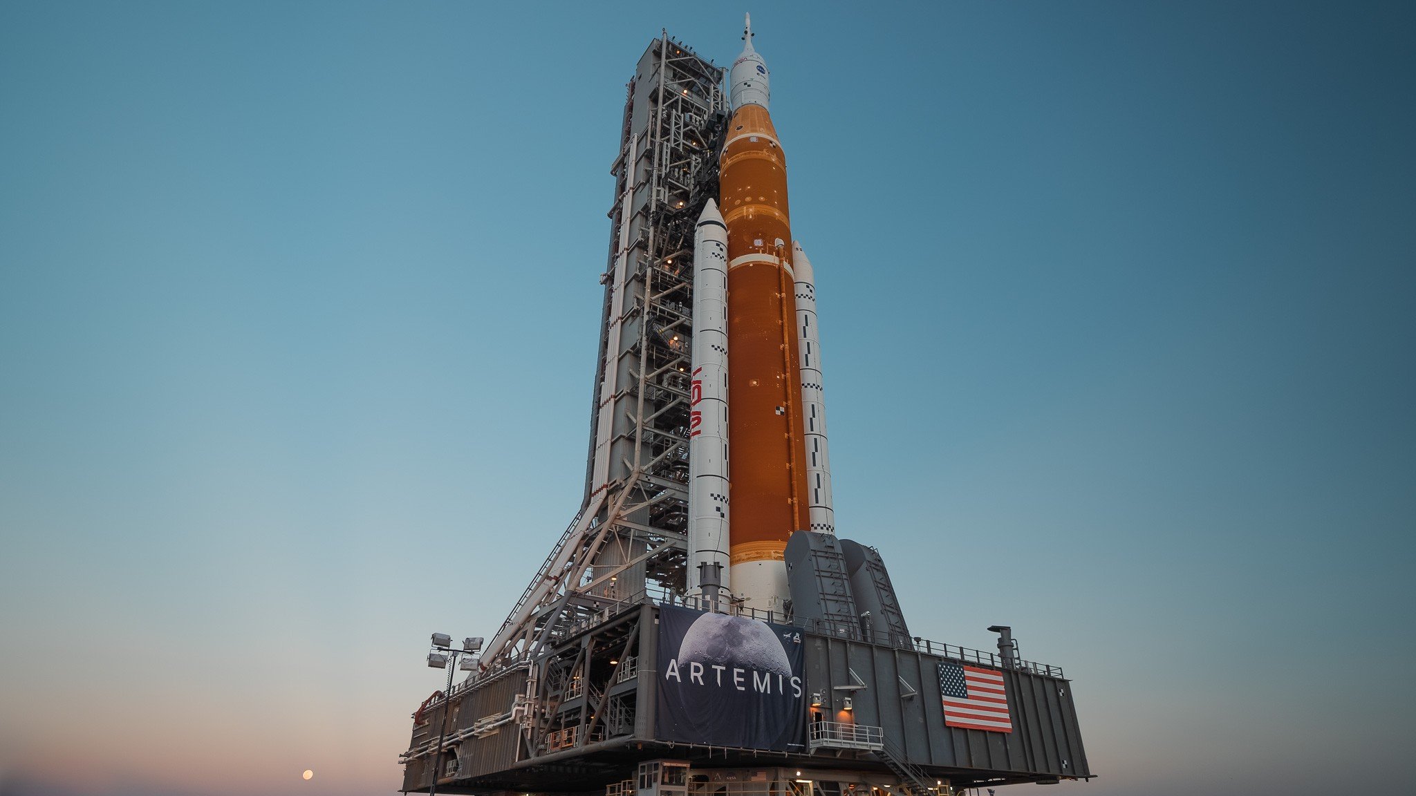 A large space rocket on a launch pad with a banner reading 'ARTEMIS' and an American flag, under a clear sky at dusk.