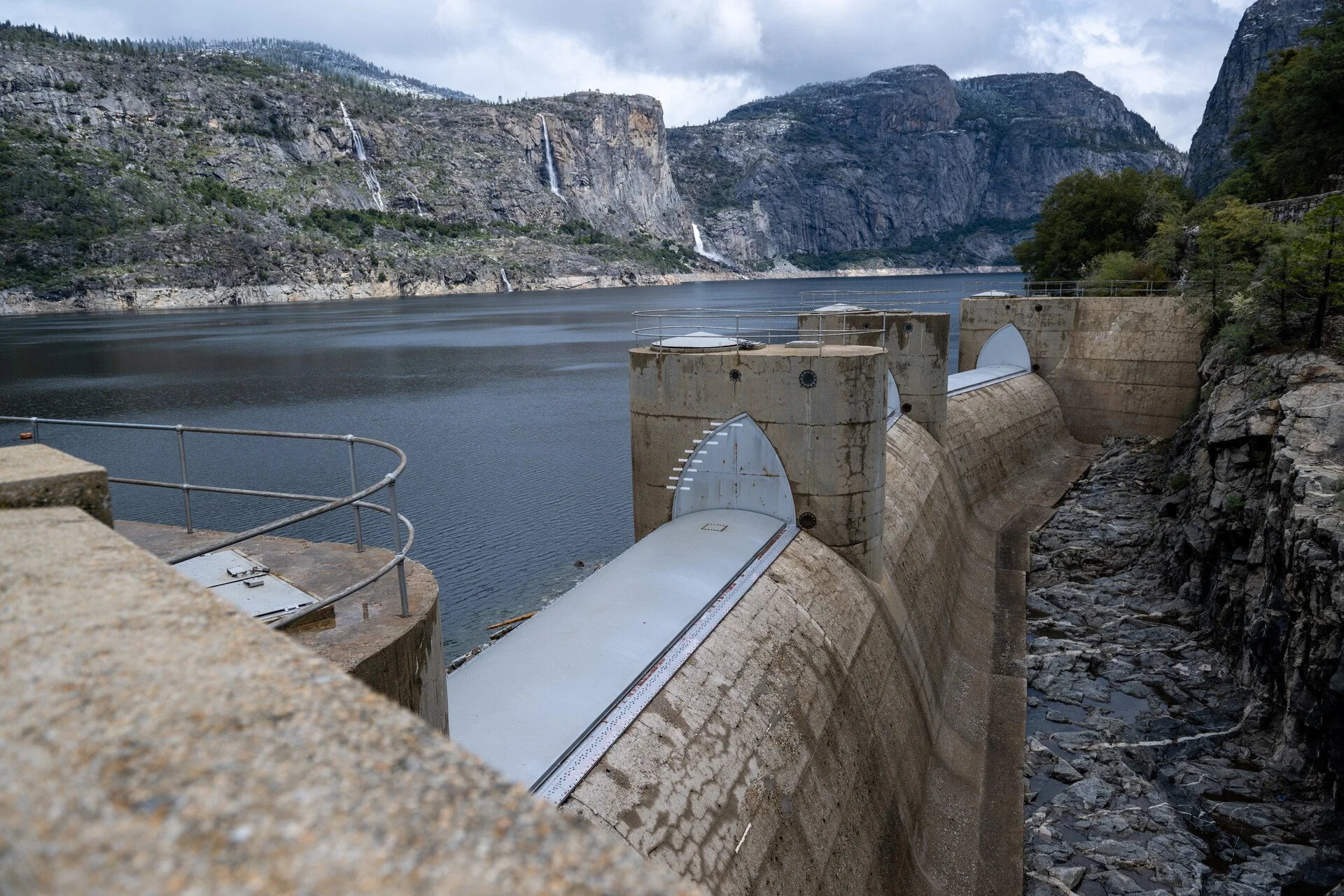 View of a dam with water release gates in a mountainous landscape with a large reservoir, rocks, trees, and waterfalls in the background under cloudy skies.