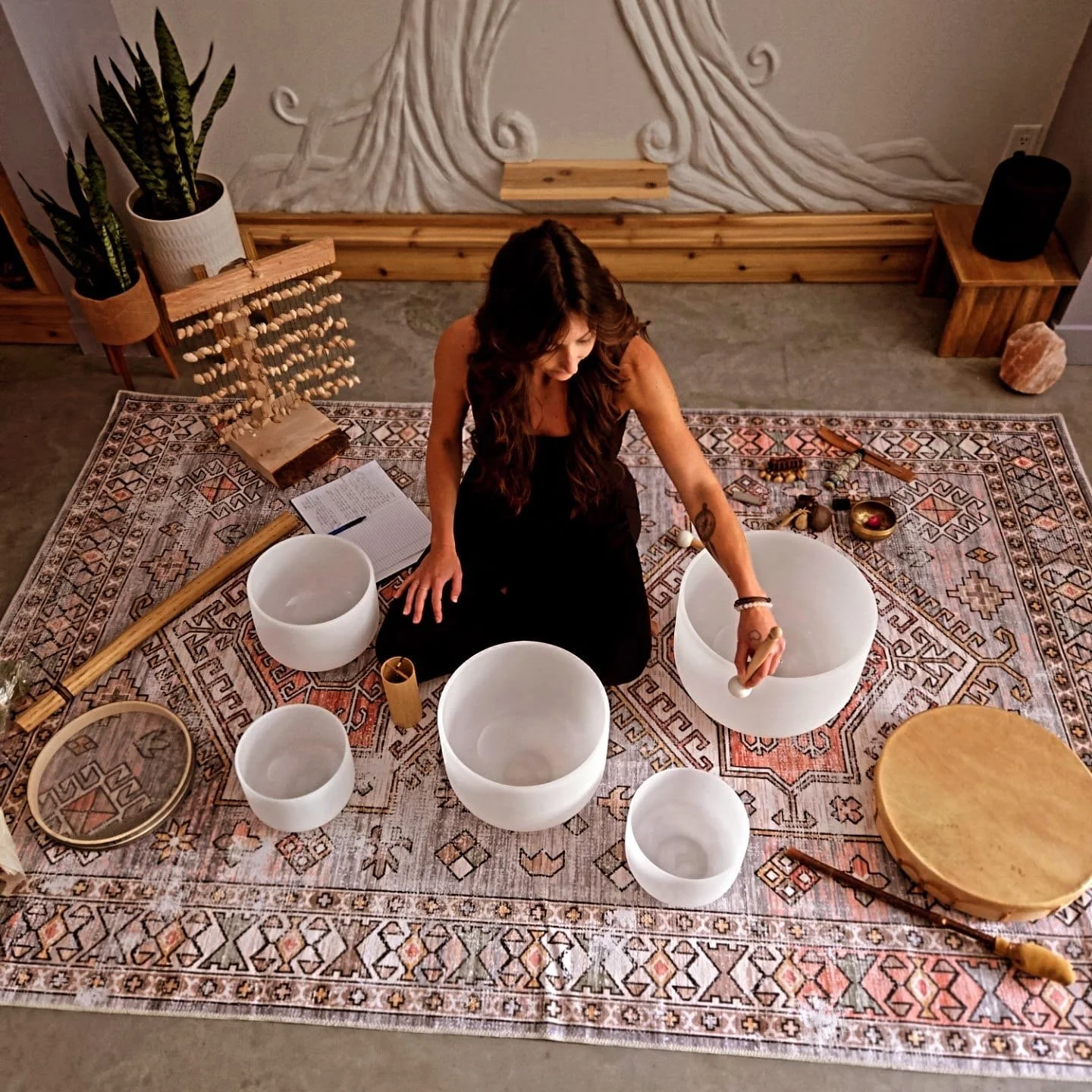 Woman sitting on a patterned rug creating singing bowls, surrounded by musical and healing instruments, with a notebook, plants, and decorative objects in a cozy room.