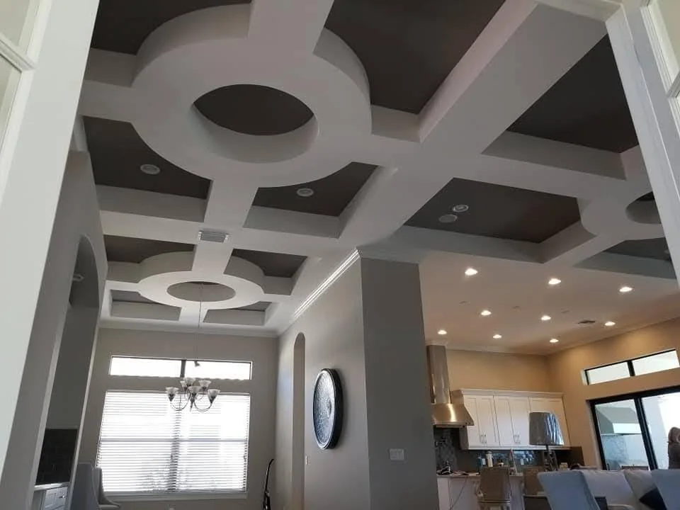 Interior view of a modern home with intricate ceiling design, large windows with blinds, a dining table with chairs, and a kitchen area with white cabinetry and stainless steel appliances.
