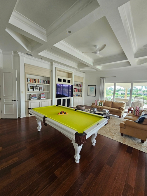 Living room with a bright yellow pool table, beige sofas, built-in white bookshelves, a wall-mounted TV, and large windows showing a backyard with trees.