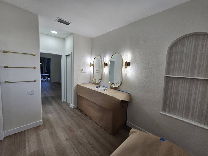 Bathroom with two oval mirrors above a beige vanity with a curved wood front, mounted wall lights, a small sink, and a window with textured curtain. Light-colored wooden flooring.