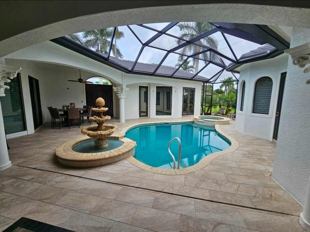 Indoor pool area with a curved swimming pool, a small fountain, and a screened enclosure with a view of palm trees outside.
