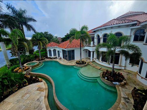 Luxurious backyard with a swimming pool, surrounded by palm trees and tropical plants, adjacent to a large white house with a red tile roof.