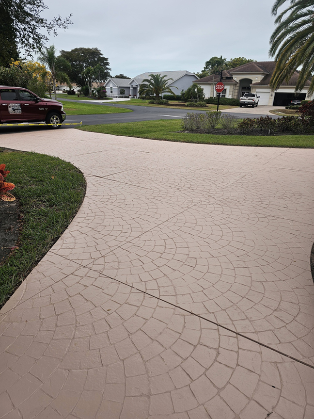 View of a suburban neighborhood with a pink stamped concrete driveway, green lawns, palm trees, and houses in the background, with a stop sign at the street corner.