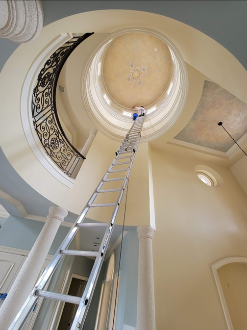 A tall ladder extends upward towards a circular, domed ceiling with decorative molding and a partly painted mural, inside a spacious foyer with light-colored walls and ornate columns.