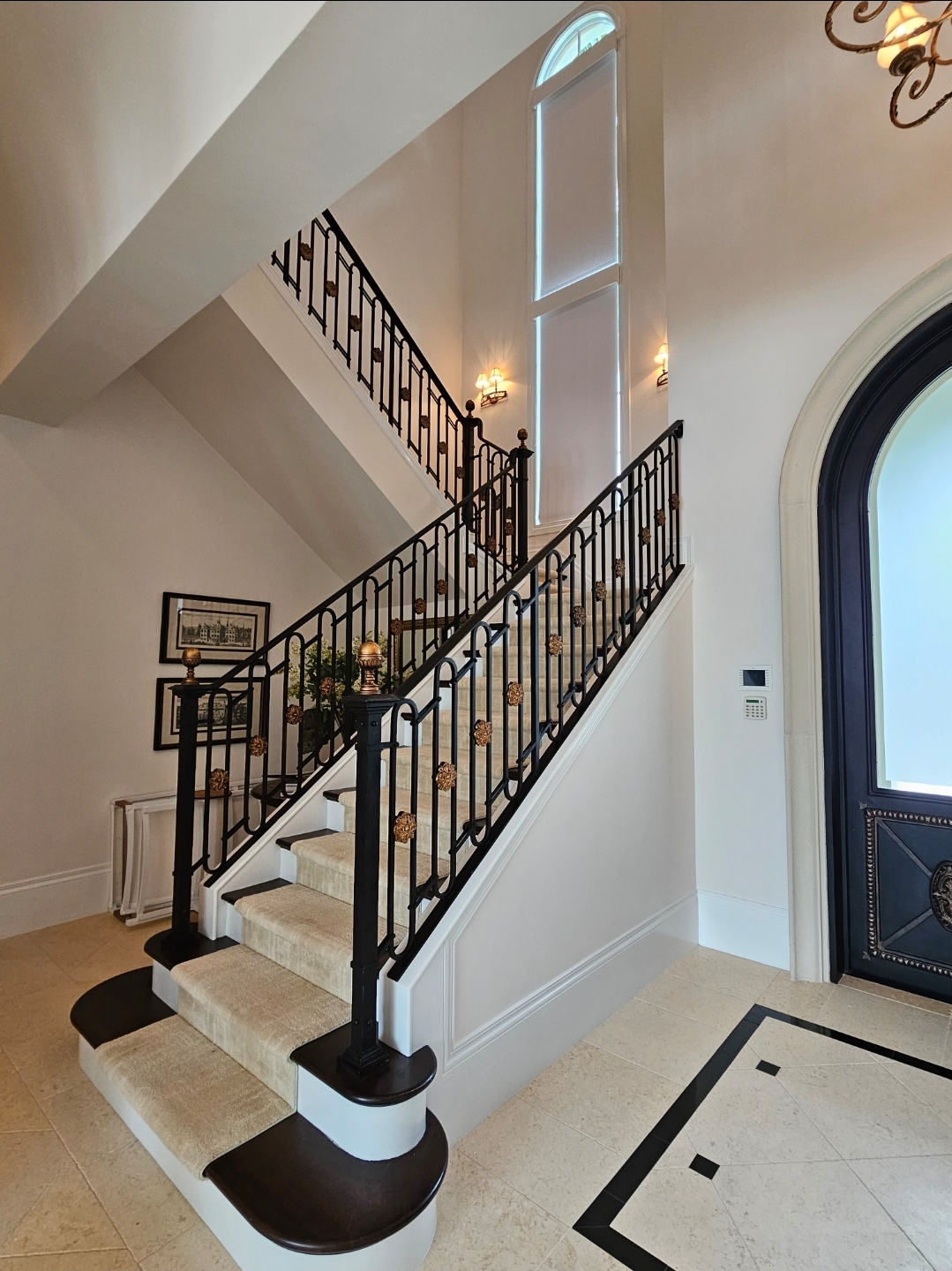 Interior of a house with a staircase featuring black metal railings with decorative gold accents, beige carpeted steps, and a high ceiling with tall windows letting in natural light. There's a door with a decorative black and gold design, wall-mounted lights, and framed artwork on the wall.
